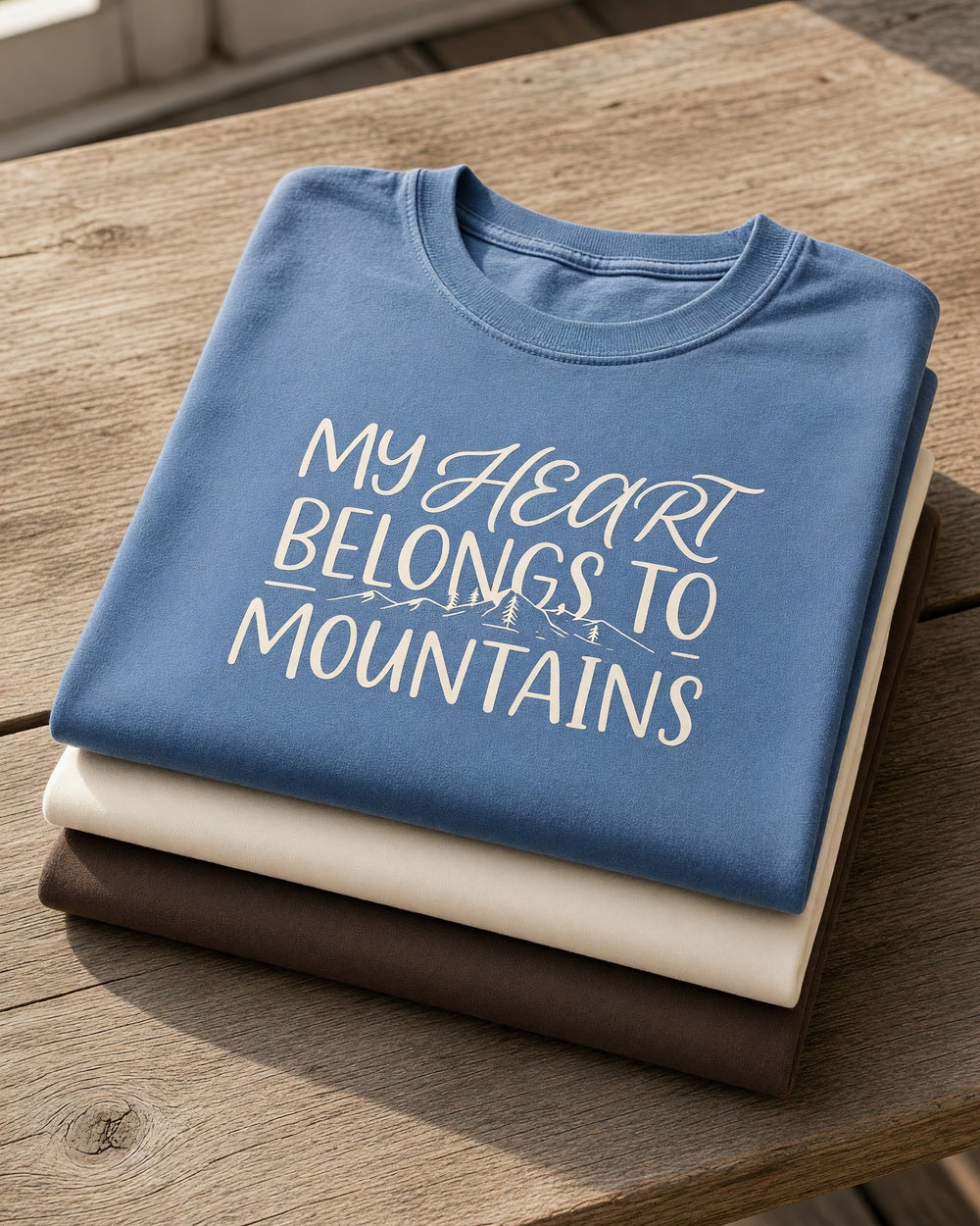 A stack of folded Comfort Colors t-shirts on a porch table, My Heart Belongs to Mountains on blue jean on top of ivory and espresso