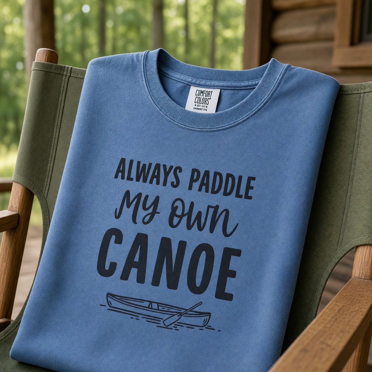 A blue jean Comfort Colors crewneck sweatshirt printed with Always Paddle My Own Canoe, draped over a camp chair on a cabin porch