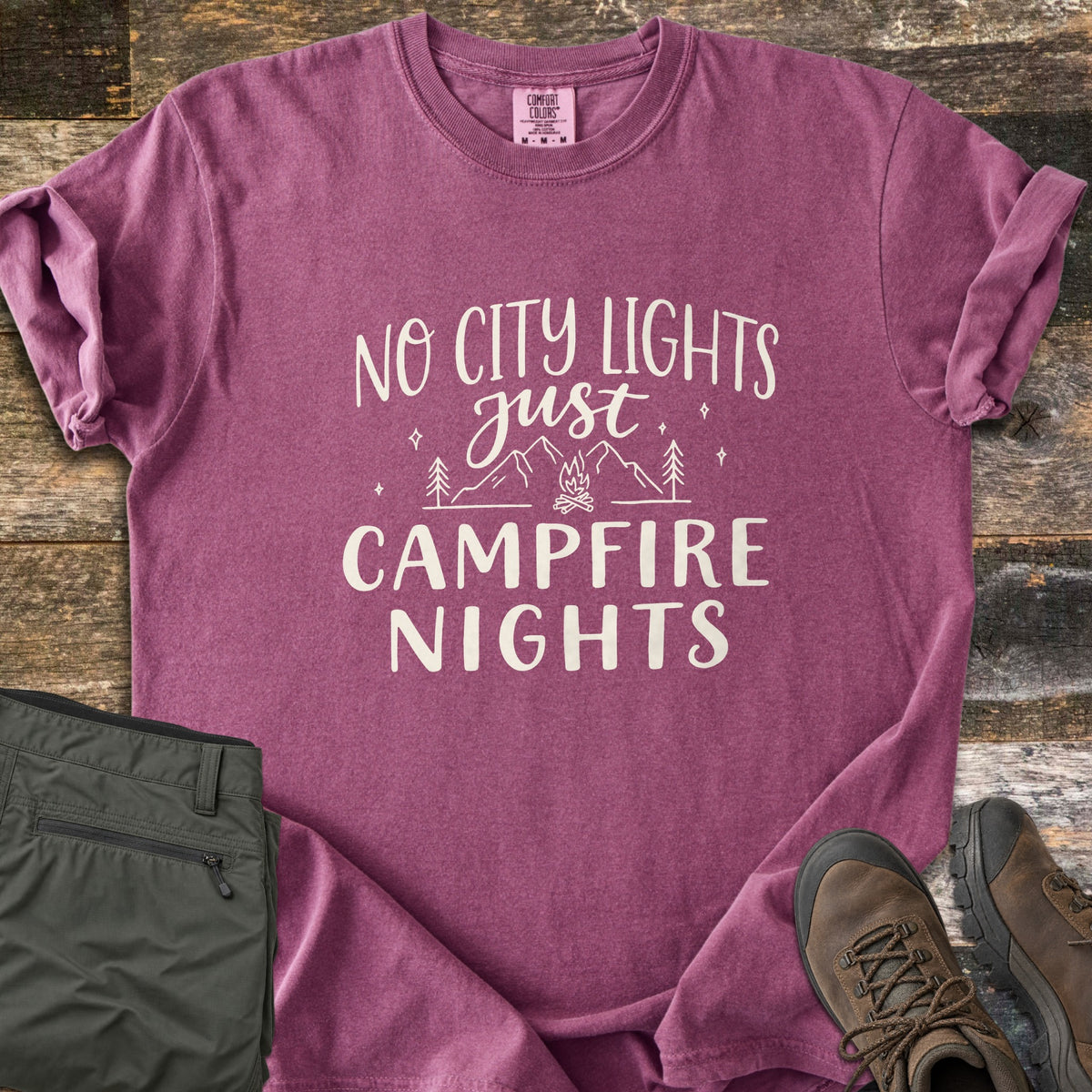 Campfire Nights Comfort Colors T-shirt in Berry, laid flat. Cream line art of mountains, pine trees, stars and a campfire sits between the words No City Lights just Campfire Nights. Sleeves rolled, on weathered wood with olive hiking pants and brown boots.