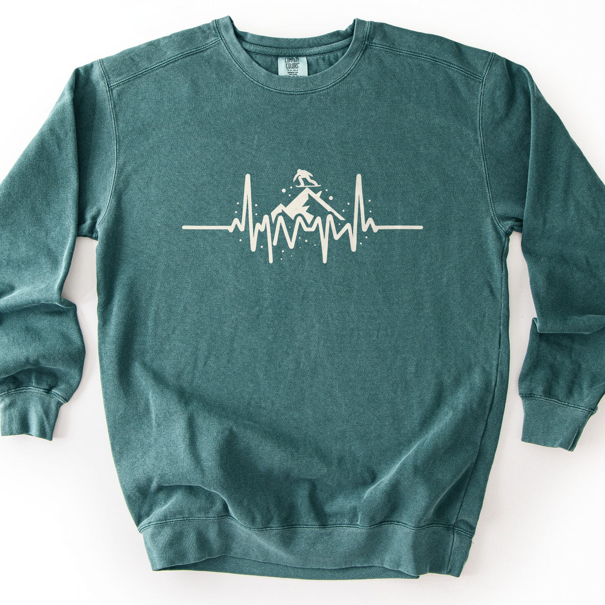 Snow Boarder Heartbeat Comfort Colors crewneck sweatshirt in Blue Spruce, laid flat. A cream heartbeat line runs across the chest, rising into a snowy mountain peak with a snowboarder riding on top and small snow dots around it. No words are printed. The sweatshirt lies on a white background.