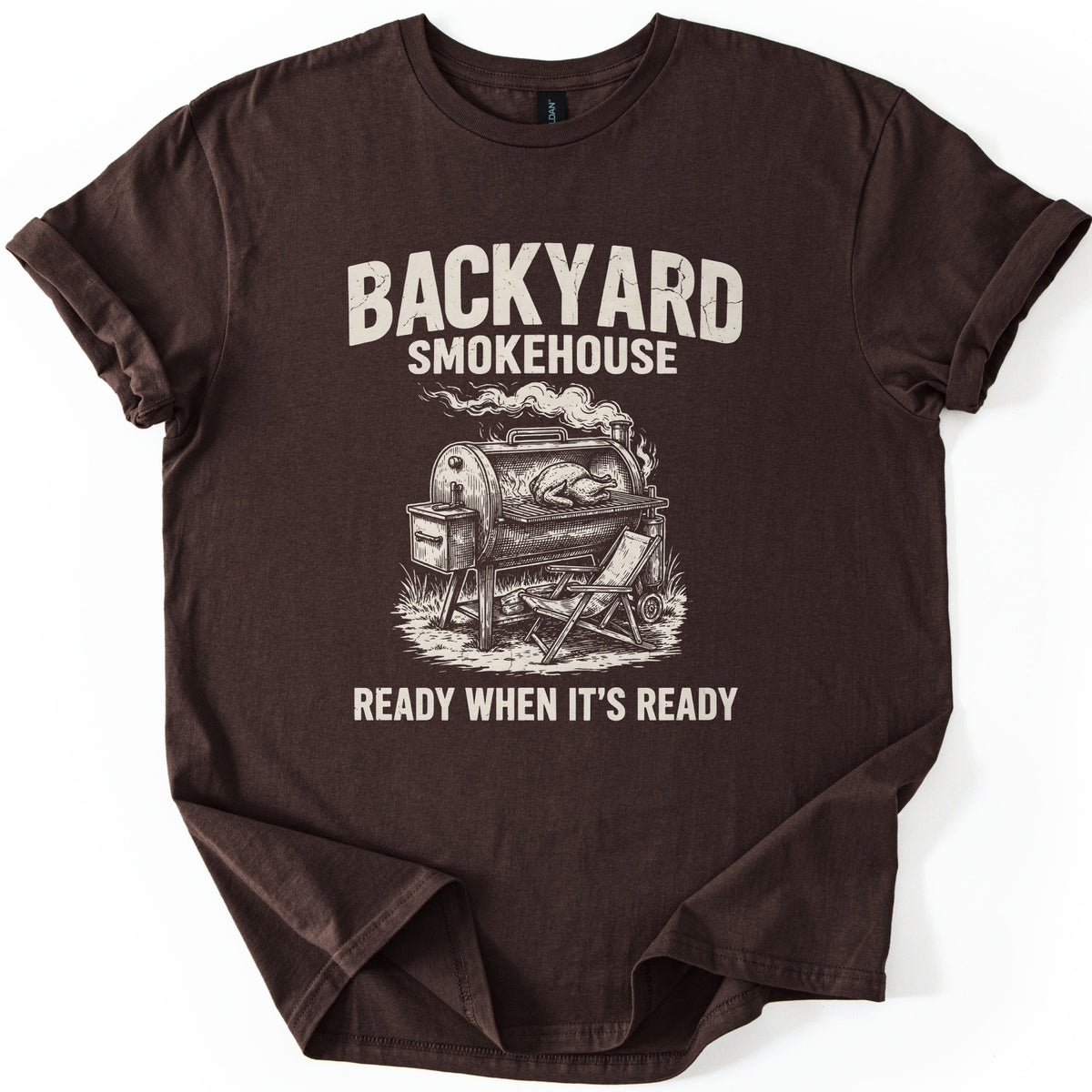 Backyard Smokehouse Comfort Colors T-shirt in Dark Chocolate, laid flat. Cream distressed letters read BACKYARD SMOKEHOUSE above a sketch of a smoking barrel smoker with a whole bird on the grate and a folding chair beside it, then READY WHEN IT'S READY. Sleeves cuffed, on white.