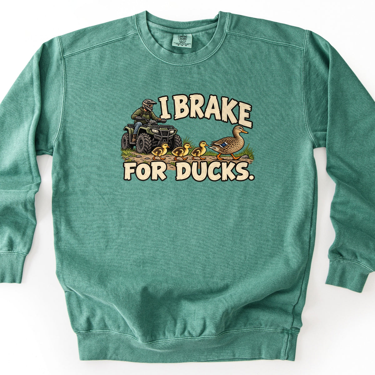 I Brake for Ducks Comfort Colors crewneck sweatshirt in Light Green, laid flat. A helmeted rider on a green ATV stops behind a mother mallard leading three yellow ducklings along a dirt path, framed by the words I Brake For Ducks. in cream letters. It lies on a white surface.
