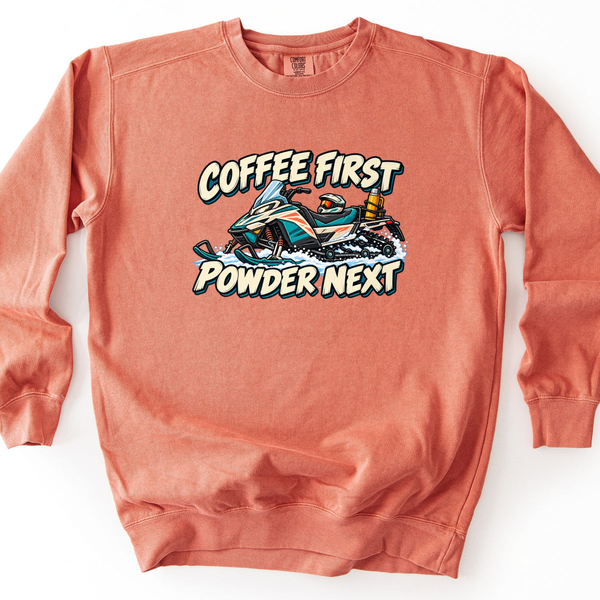 Coffee First Powder Next Comfort Colors crewneck sweatshirt in Terracotta, laid flat. A cartoon teal and white snowmobile with a helmet on the seat and a yellow thermos strapped to the back kicks up snow between the words Coffee First and Powder Next. Laid on a white background.