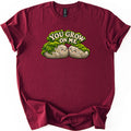 You Grow on Me Comfort Colors T-shirt in Maroon, laid flat. Two smiling cartoon rocks with rosy cheeks lean cheek to cheek under a blanket of green moss, beneath the arched words You Grow On Me. The shirt lies on a white surface with its sleeves cuffed.