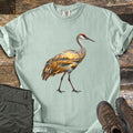 Crane Marsh Sunrise Comfort Colors T-shirt in Bay, laid flat. A sandhill crane with a red crown walks across the chest, its body filled with a sunrise marsh scene of golden reeds and a winding stream. Rolled sleeves; gray pants and brown hiking boots sit on weathered wood.