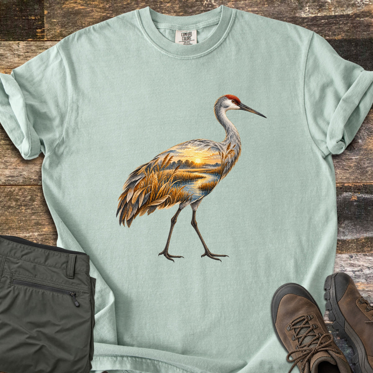 Crane Marsh Sunrise Comfort Colors T-shirt in Bay, laid flat. A sandhill crane with a red crown walks across the chest, its body filled with a sunrise marsh scene of golden reeds and a winding stream. Rolled sleeves; gray pants and brown hiking boots sit on weathered wood.