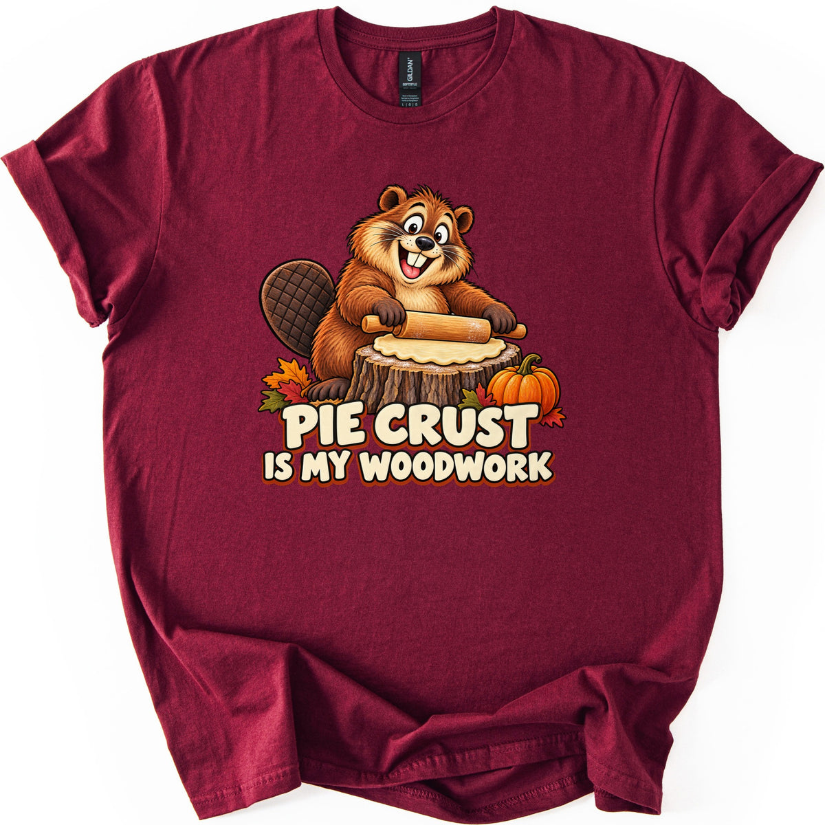 Pie Crust Is My Woodwork Comfort Colors T-shirt in Maroon, laid flat. A grinning cartoon beaver rolls pie dough with a rolling pin on a tree stump, beside a small pumpkin and autumn leaves, above the words Pie Crust Is My Woodwork. Cuffed sleeves, on a white background.
