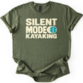 Silent Mode Comfort Colors T-shirt in Military Green, laid flat. Bold cream block letters read Silent Mode Kayaking, with a teal circle showing a kayak and paddle from above beside the word Mode. The shirt has rolled sleeves on a white background.