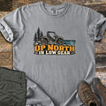 Up North in Low Gear Comfort Colors T-shirt in Grey, laid flat. A side-by-side off-road vehicle sits on rocks by pine trees and a lake above the words Up North in Low Gear. Olive cargo pants and brown hiking boots lie beside it on rustic wood planks.