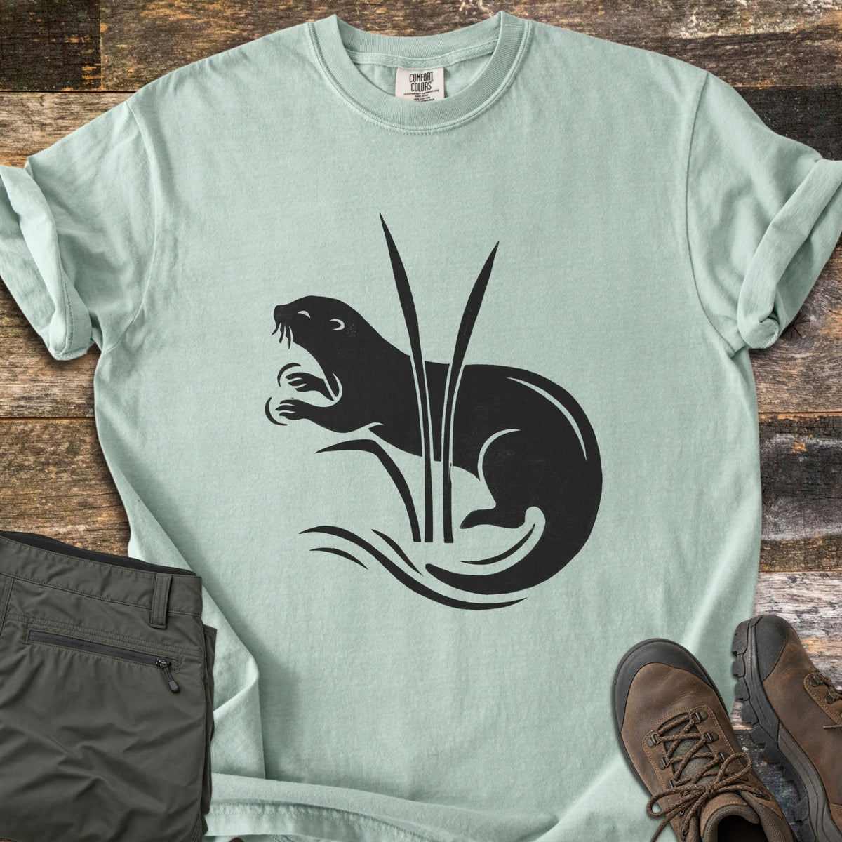 Otter In The Reeds Comfort Colors T-shirt in Bay, laid flat. A black silhouette of an otter with open mouth and raised paws curls behind two tall reeds above wavy water lines, no words. Folded olive hiking pants and brown hiking boots lie on weathered wood.