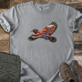 Red Canyon Raven Comfort Colors T-shirt in Grey, laid flat. A black raven in flight, its body and wings filled with red canyon cliffs and a winding blue river. Sleeves rolled, on weathered wood with folded olive hiking pants and brown hiking boots.