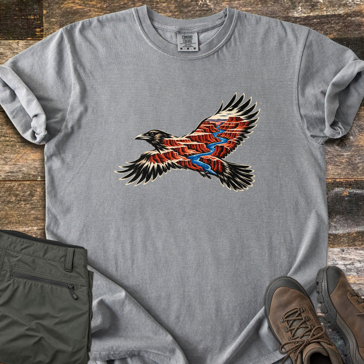 Red Canyon Raven Comfort Colors T-shirt in Grey, laid flat. A black raven in flight, its body and wings filled with red canyon cliffs and a winding blue river. Sleeves rolled, on weathered wood with folded olive hiking pants and brown hiking boots.