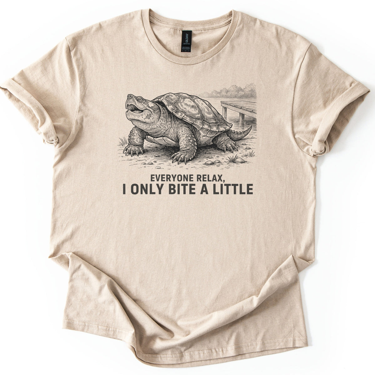 Everyone Relax I Only Bite A Little Comfort Colors T-shirt in Sand, laid flat. A black ink drawing of a snapping turtle with its mouth open on a shoreline by a wooden dock, above the words Everyone Relax, I Only Bite A Little. Sleeves are rolled on a white background.