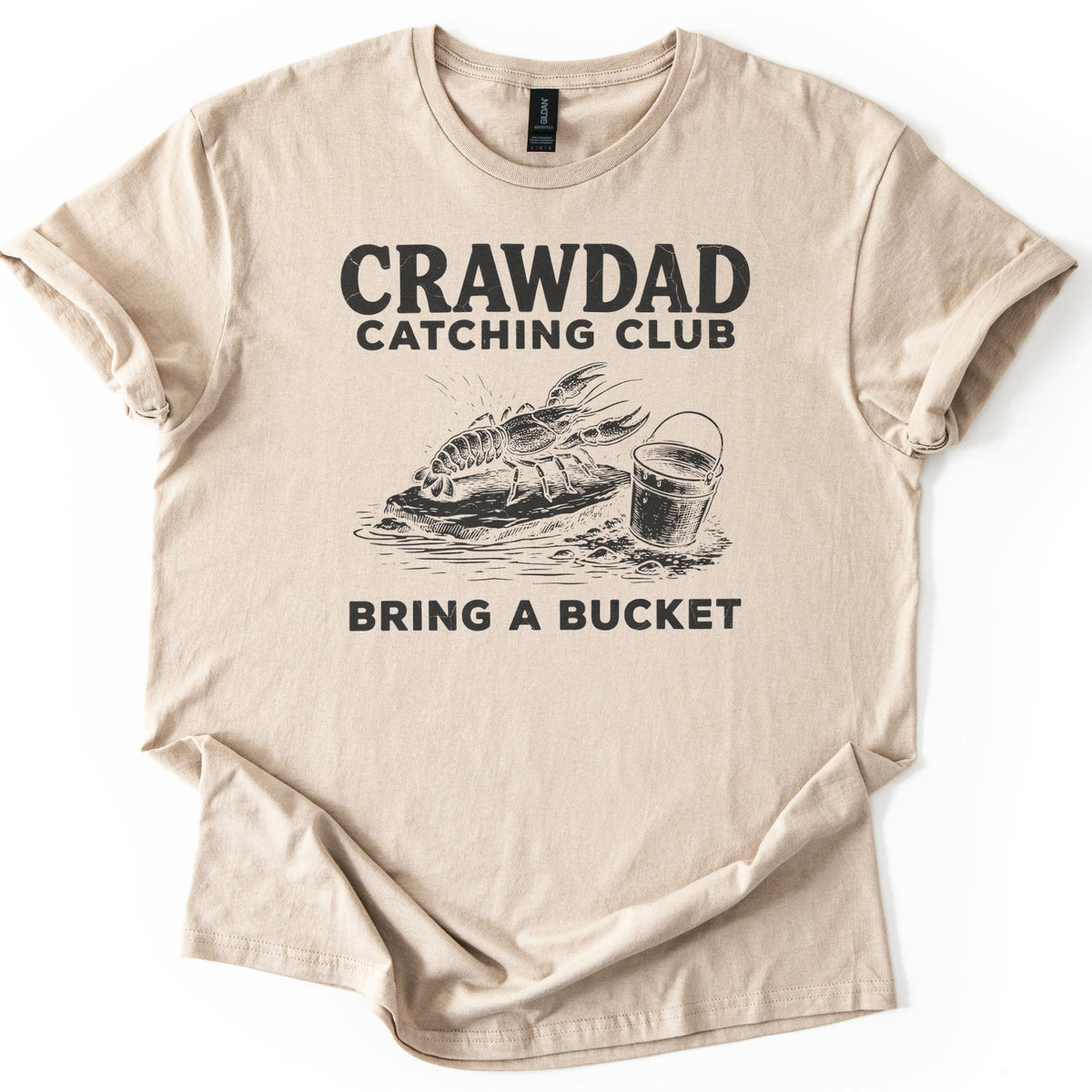 Crawdad Catching Club Comfort Colors T-shirt in Sand, laid flat. Black print reads Crawdad Catching Club above a sketched crawdad on a flat rock at the water's edge beside a metal bucket, with Bring a Bucket below. Cuffed sleeves on a white background.