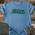 Helice Comfort Colors T-shirt in Blue Jean, laid flat. The word Helice is printed in dark green script lettering with a swash underline across the chest. Sleeves are rolled, on a rustic wood floor beside folded olive cargo pants and brown leather hiking boots.