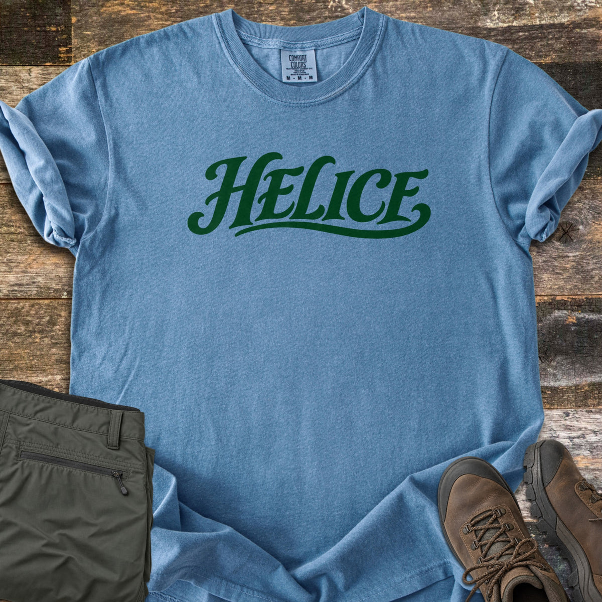 Helice Comfort Colors T-shirt in Blue Jean, laid flat. The word Helice is printed in dark green script lettering with a swash underline across the chest. Sleeves are rolled, on a rustic wood floor beside folded olive cargo pants and brown leather hiking boots.