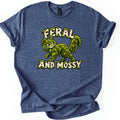Feral and Mossy Comfort Colors T-shirt in Heather Navy, laid flat. A smiling cartoon cat made of dripping green moss with brown twigs showing through, between moss-covered cream letters reading Feral and Mossy. The shirt lies on a white background with cuffed sleeves.