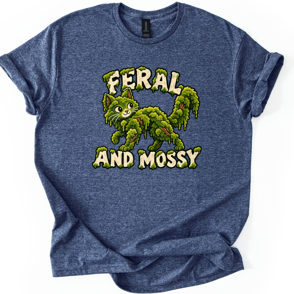 Feral and Mossy Comfort Colors T-shirt in Heather Navy, laid flat. A smiling cartoon cat made of dripping green moss with brown twigs showing through, between moss-covered cream letters reading Feral and Mossy. The shirt lies on a white background with cuffed sleeves.