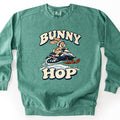 Bunny Hop Comfort Colors crewneck sweatshirt in Light Green, laid flat. A grinning cartoon bunny in a white helmet rides a navy, white and orange snowmobile over a snowbank between the words Bunny Hop in cream bubble letters. The sweatshirt lies on a white surface.