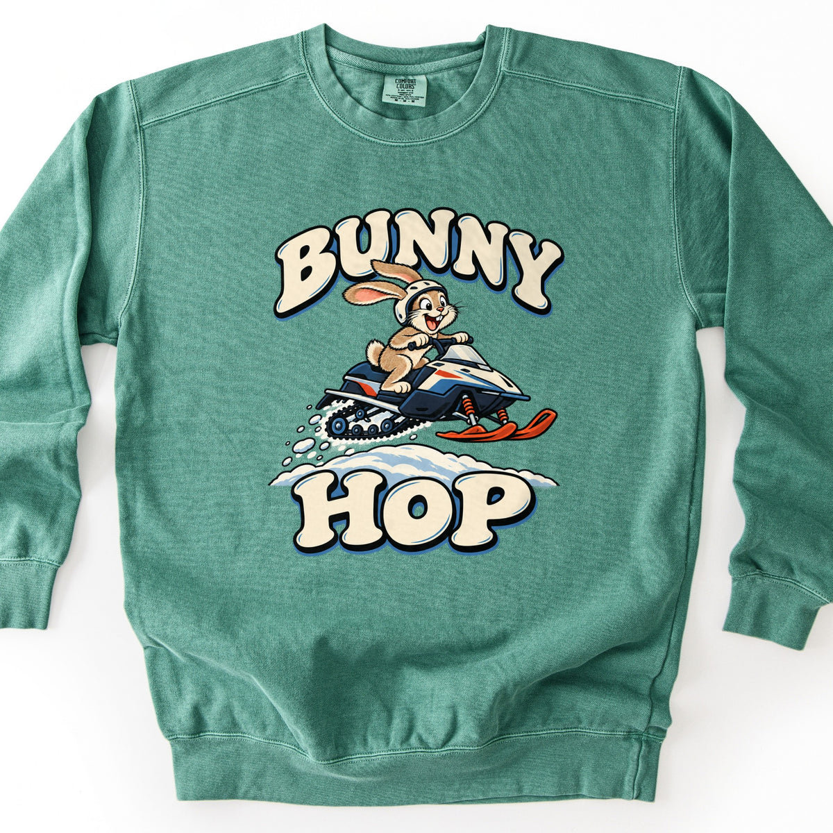 Bunny Hop Comfort Colors crewneck sweatshirt in Light Green, laid flat. A grinning cartoon bunny in a white helmet rides a navy, white and orange snowmobile over a snowbank between the words Bunny Hop in cream bubble letters. The sweatshirt lies on a white surface.