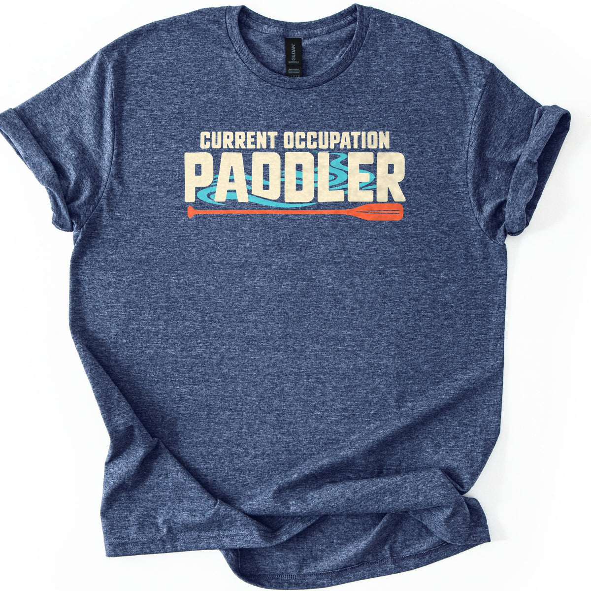 Current Occupation Paddler Comfort Colors T-shirt in Heather Navy, laid flat. The words Current Occupation above a large Paddler in cream letters over swirling turquoise water, with an orange canoe paddle underneath. The shirt has rolled sleeves and lies on a white background.