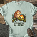 The Night Shift Has Spores Comfort Colors T-shirt in Bay, laid flat. A sleepy brown owl sits on a mossy branch beside glowing orange mushrooms, ferns and red berries under small stars, above The Night Shift Has Spores in dark green. Olive pants and brown hiking boots lie on weathered wood.