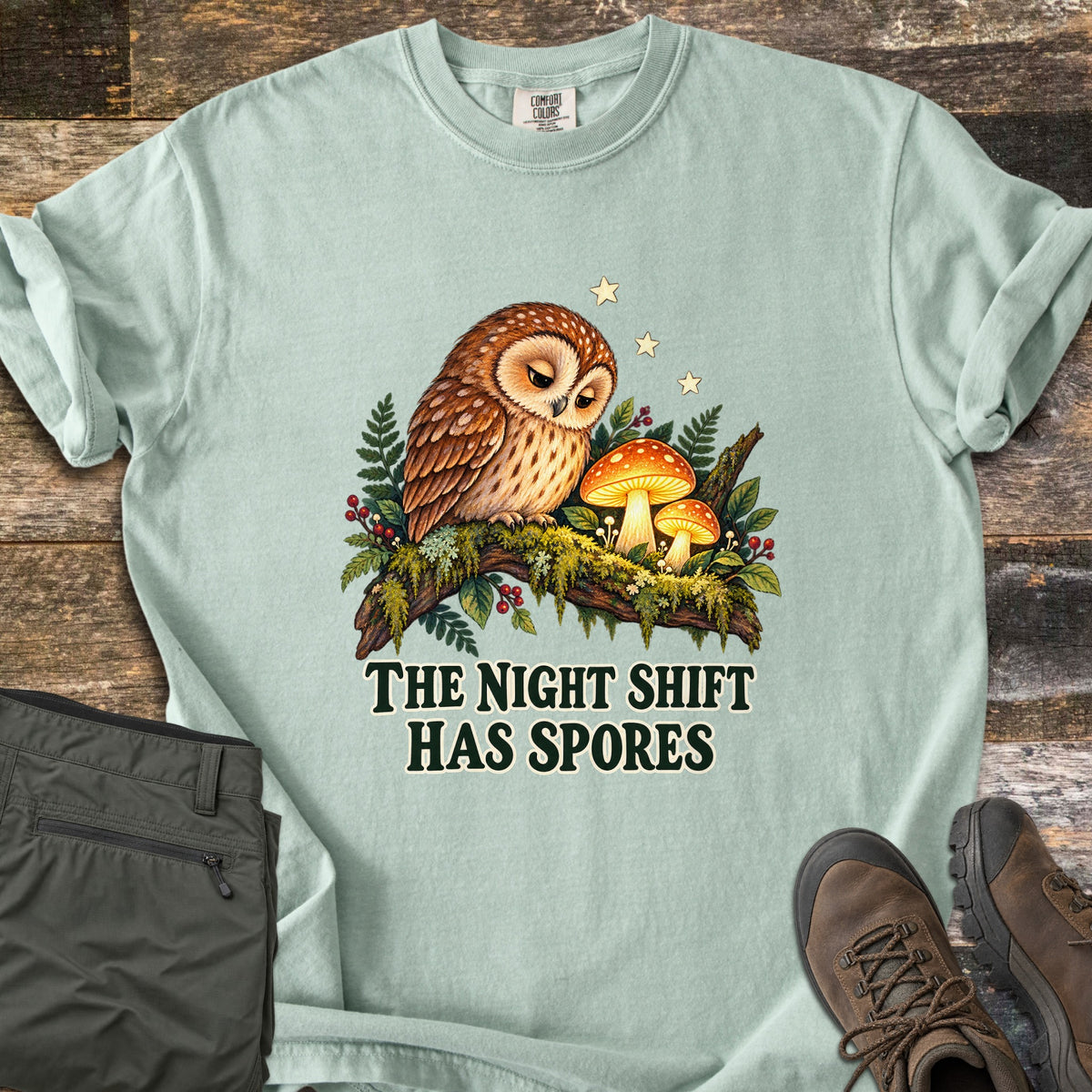 The Night Shift Has Spores Comfort Colors T-shirt in Bay, laid flat. A sleepy brown owl sits on a mossy branch beside glowing orange mushrooms, ferns and red berries under small stars, above The Night Shift Has Spores in dark green. Olive pants and brown hiking boots lie on weathered wood.