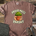 Proud Moss Parent Comfort Colors T-shirt in Espresso, laid flat. A cartoon hand holds a terracotta pot of smiling green moss between the words Proud Moss Parent. Sleeves are rolled, with green hiking pants and brown hiking boots on a weathered wood background.