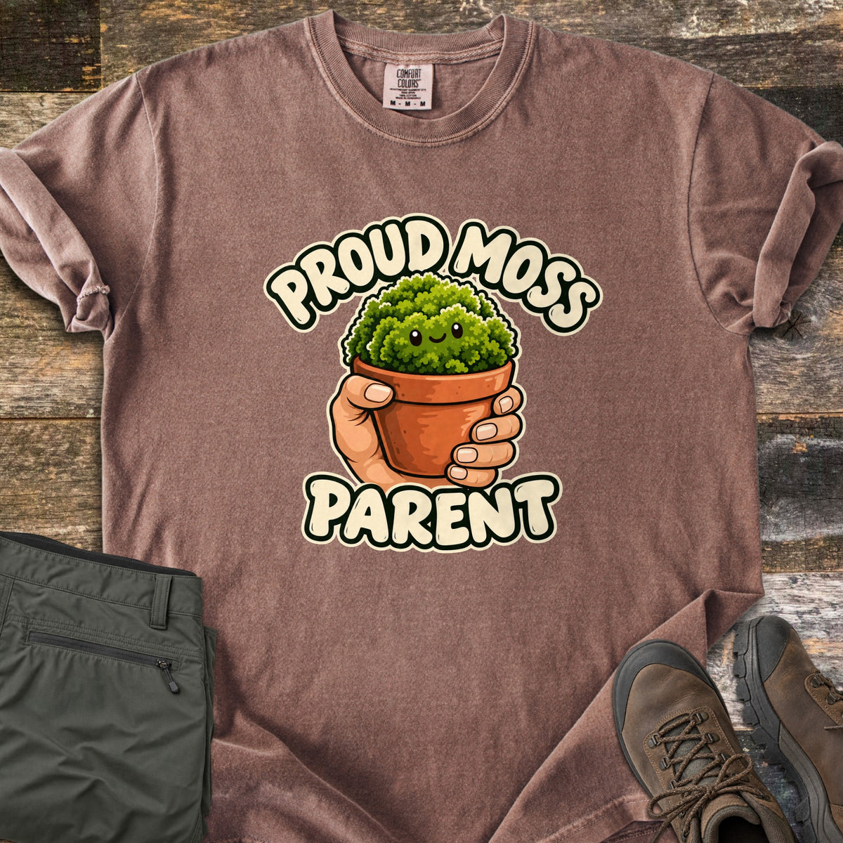 Proud Moss Parent Comfort Colors T-shirt in Espresso, laid flat. A cartoon hand holds a terracotta pot of smiling green moss between the words Proud Moss Parent. Sleeves are rolled, with green hiking pants and brown hiking boots on a weathered wood background.