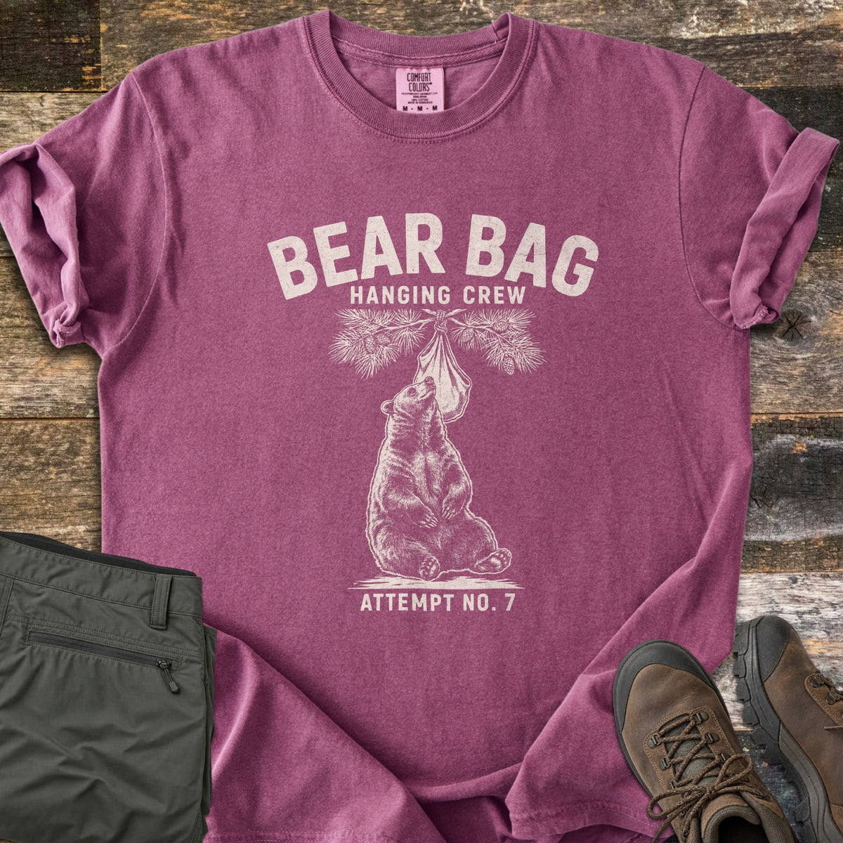 Bear Bag Hanging Crew Comfort Colors T-shirt in Berry, laid flat. A sketched bear sits under a pine branch sniffing a hanging food bag, with the words Bear Bag Hanging Crew above and Attempt No. 7 below. Olive hiking pants and brown hiking boots lie beside it on weathered wood.