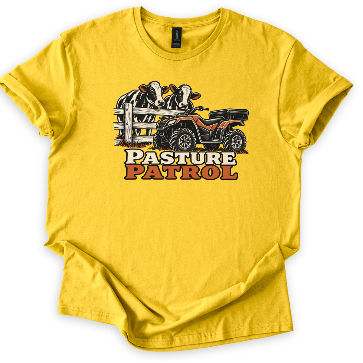 Pasture Patrol Comfort Colors T-shirt in Daisy, laid flat. Two black and white cows look over a wooden fence beside an orange ATV with a rear cargo box, above the words Pasture Patrol. The shirt lies on a white background with its sleeves rolled.