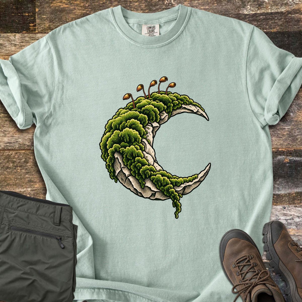 Moss Moon Comfort Colors T-shirt in Bay, laid flat. A pale stone crescent moon draped in thick green moss, with thin brown spore stalks rising from the top. Sleeves rolled, on weathered wood planks beside folded olive hiking pants and brown hiking boots.