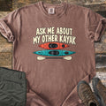 Other Kayak Comfort Colors T-shirt in Espresso, laid flat. The words Ask Me About My Other Kayak in cream letters above an orange kayak, a teal kayak, and a cream paddle. Sleeves rolled, on weathered wood with folded olive pants and brown hiking boots.