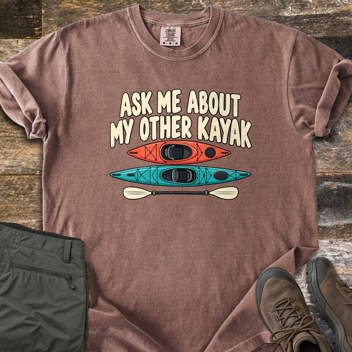 Other Kayak Comfort Colors T-shirt in Espresso, laid flat. The words Ask Me About My Other Kayak in cream letters above an orange kayak, a teal kayak, and a cream paddle. Sleeves rolled, on weathered wood with folded olive pants and brown hiking boots.