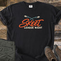 Skeet League Night Comfort Colors T-shirt in Black, laid flat. Two clay targets fly in crossing blue arcs above the orange script word Skeet and cream text League Night. Olive cargo pants and brown hiking boots sit beside it on weathered wood planks.