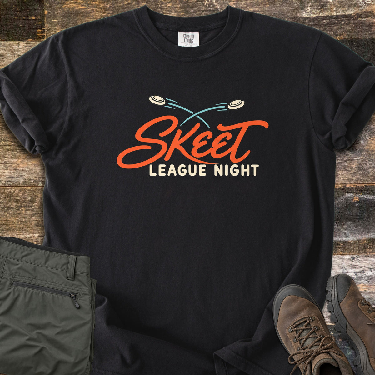 Skeet League Night Comfort Colors T-shirt in Black, laid flat. Two clay targets fly in crossing blue arcs above the orange script word Skeet and cream text League Night. Olive cargo pants and brown hiking boots sit beside it on weathered wood planks.