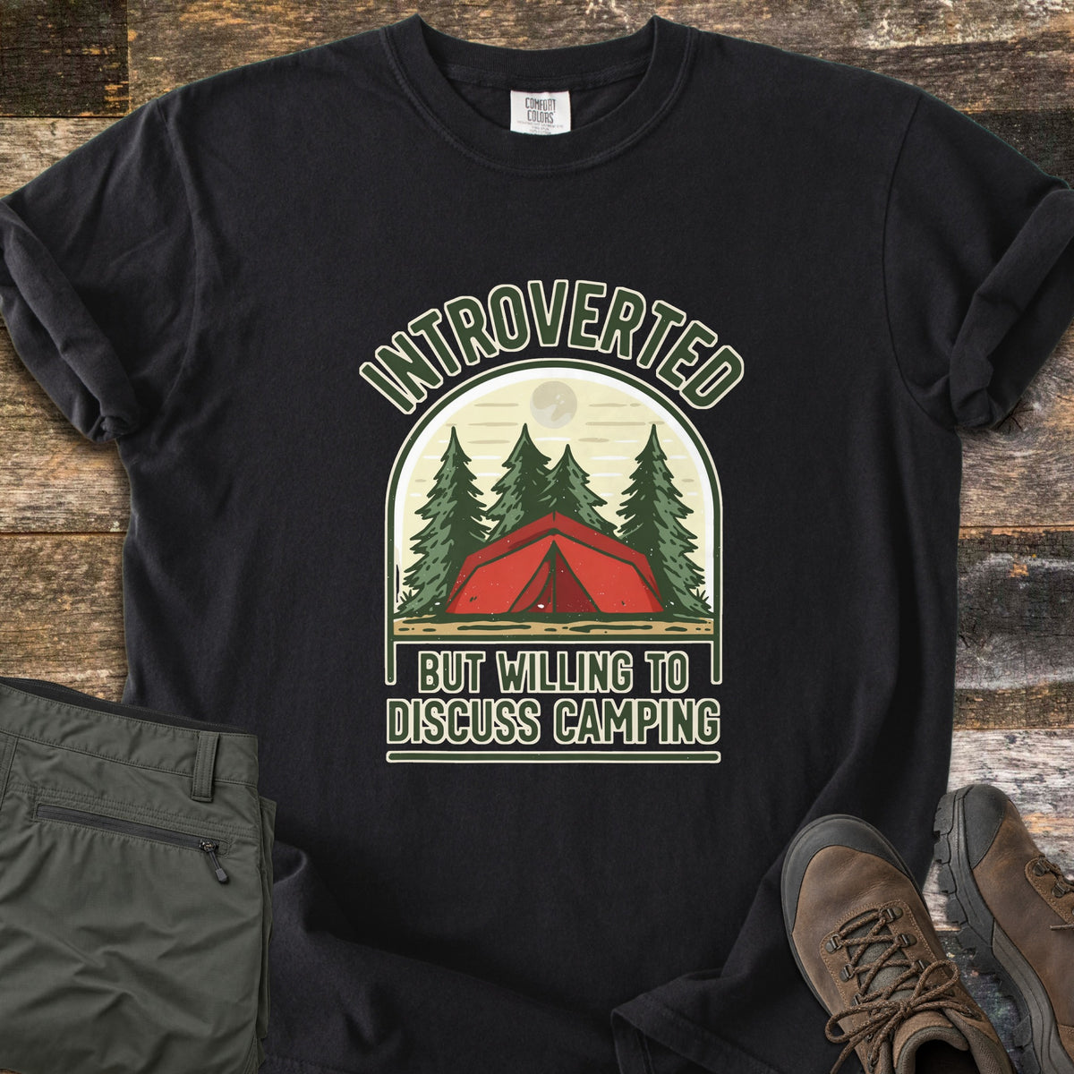 Introverted Camping Comfort Colors T-shirt in Black, laid flat. An arch shows a red tent among pine trees under a pale moon, with the words Introverted above and But Willing To Discuss Camping below. Olive pants and brown hiking boots sit beside it on weathered wood.