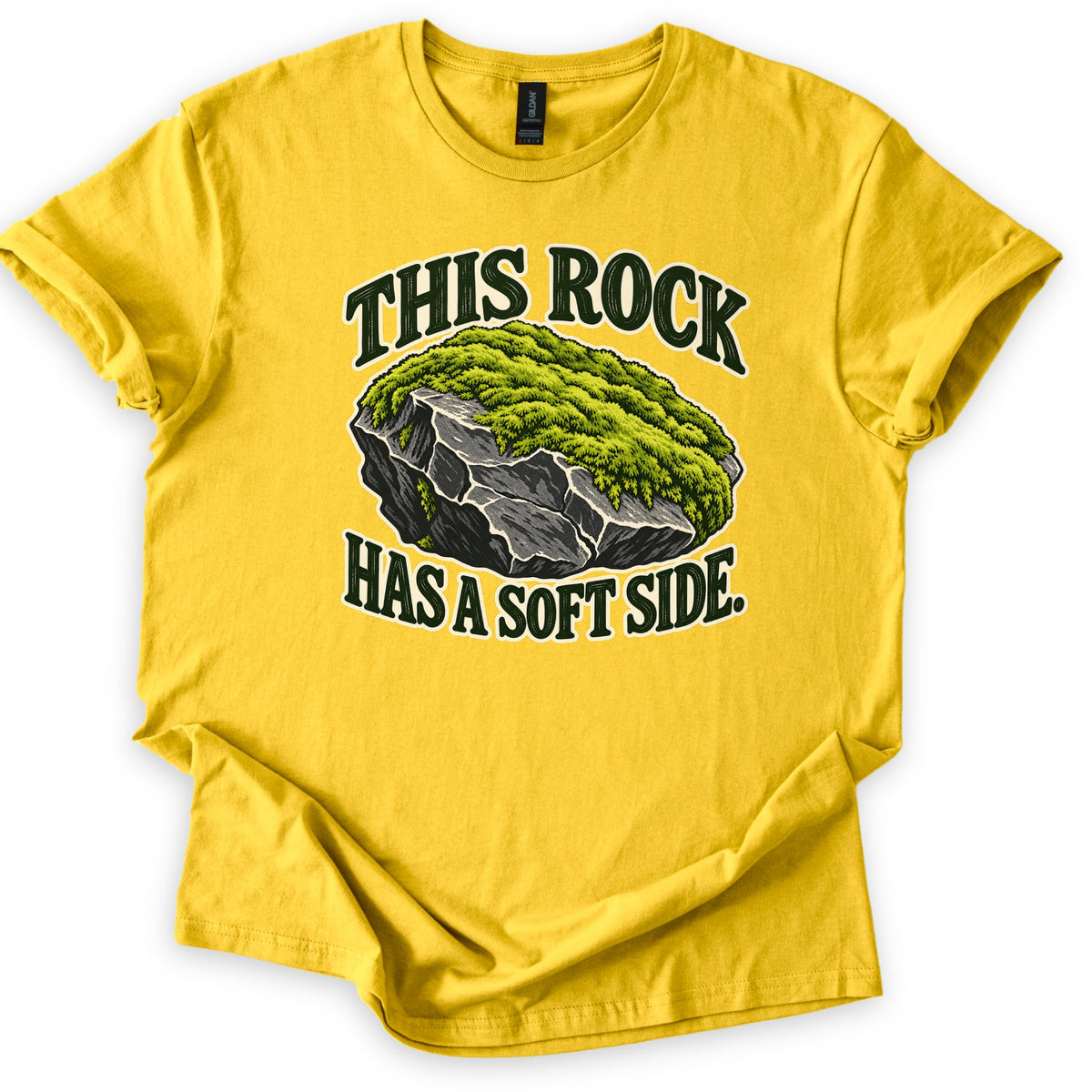 This Rock Has a Soft Side Comfort Colors T-shirt in Daisy, laid flat. A gray boulder topped with thick green moss sits between the words This Rock above and Has a Soft Side. below in dark green letters with a cream outline. The shirt lies on white with cuffed sleeves.