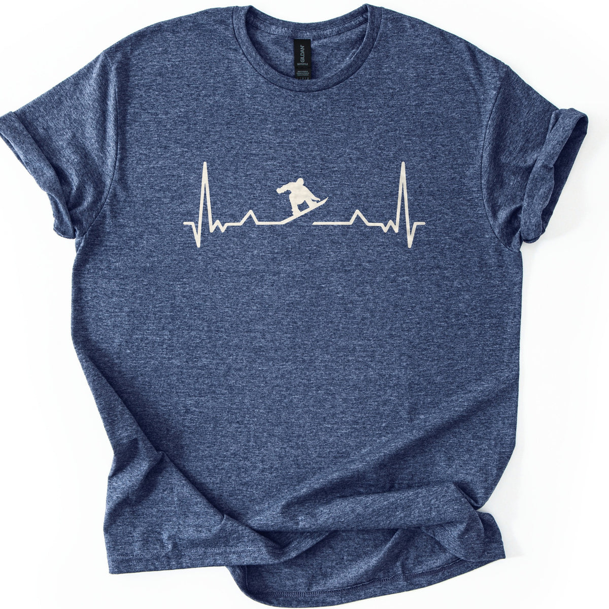 Snow Boarder Pulse II Comfort Colors T-shirt in Heather Navy, laid flat. A cream heartbeat line runs across the chest, with a snowboarder silhouette riding a dip in the line; there are no words. The shirt lies on a white surface with its sleeves rolled.