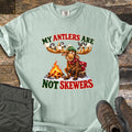 My Antlers Are Not Skewers Comfort Colors T-shirt in Bay, laid flat. A cartoon moose in a green knit hat and red scarf sits on a stump by a campfire as chickadees on its antlers hold marshmallows, with the words My Antlers Are Not Skewers. Olive pants and brown boots lie on weathered wood.