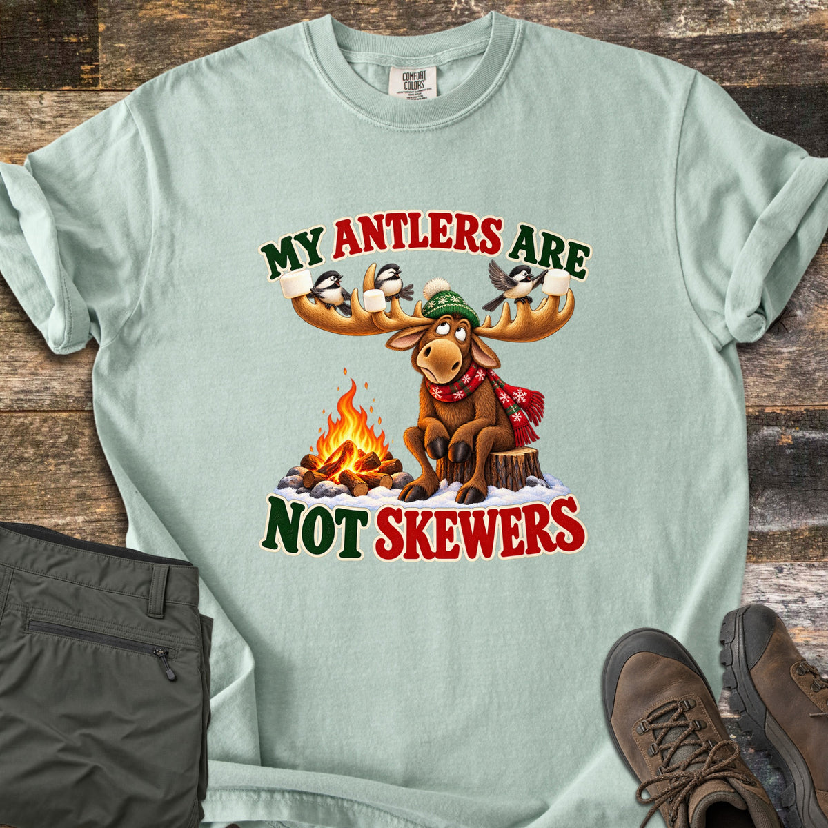 My Antlers Are Not Skewers Comfort Colors T-shirt in Bay, laid flat. A cartoon moose in a green knit hat and red scarf sits on a stump by a campfire as chickadees on its antlers hold marshmallows, with the words My Antlers Are Not Skewers. Olive pants and brown boots lie on weathered wood.