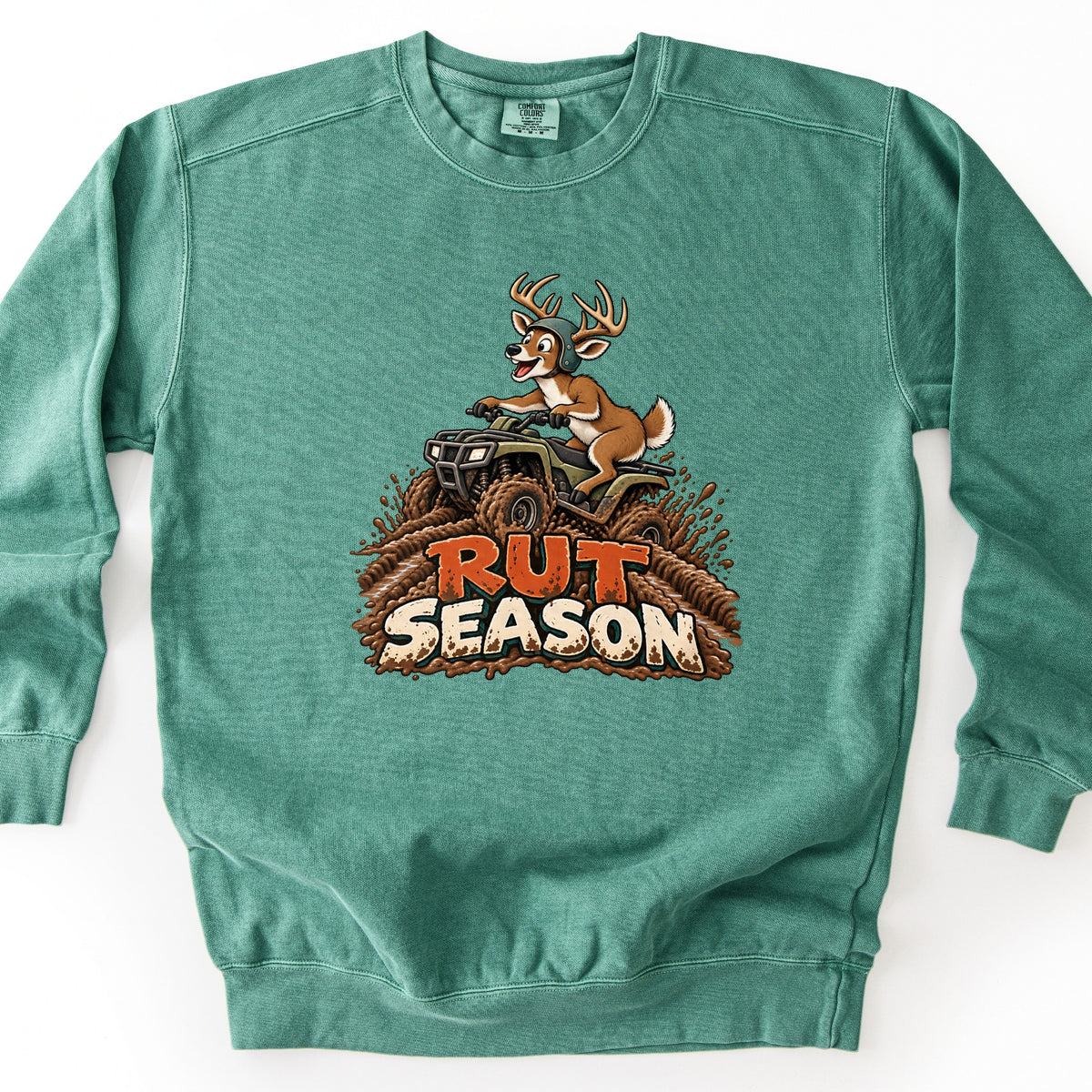 Rut Season Comfort Colors crewneck sweatshirt in Light Green, laid flat. A cartoon deer buck wearing a helmet rides a green ATV through splashing mud above the words Rut Season in orange and cream lettering. The sweatshirt lies on a white background.