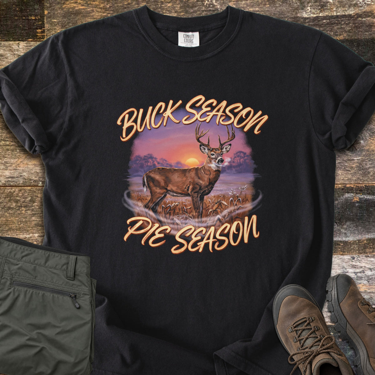 Buck Season Pie Season Comfort Colors T-shirt in Black, laid flat. A whitetail buck stands in a frosty cornfield at a purple and orange sunset between the words Buck Season and Pie Season. It lies on weathered wood beside folded olive hiking pants and brown hiking boots.
