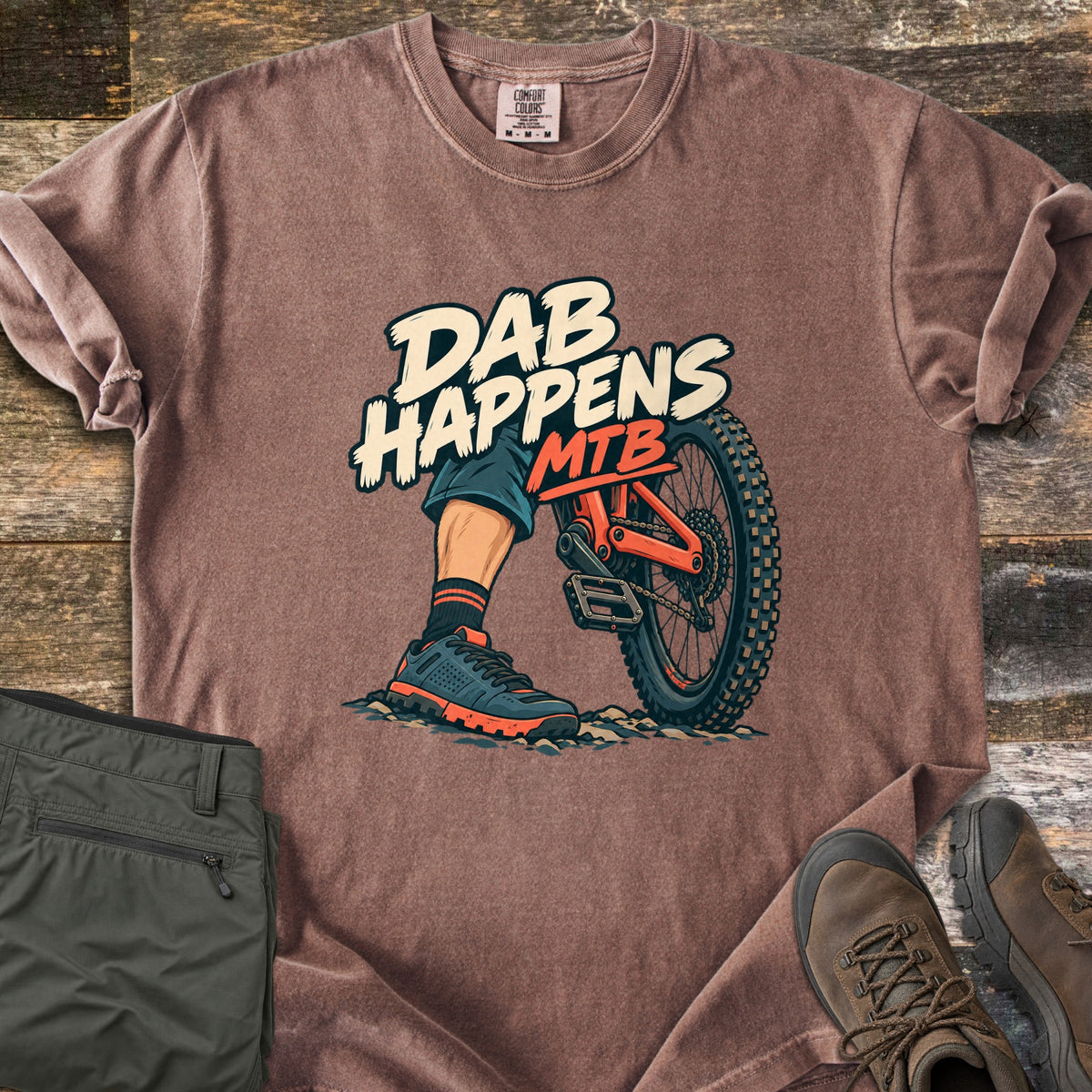 Dab Happens Comfort Colors T-shirt in Espresso, laid flat. A cartoon rider's foot in a sneaker is planted on rocky dirt beside an orange mountain bike's rear wheel, under the words Dab Happens MTB. Olive pants and brown hiking boots sit on a weathered wood surface.