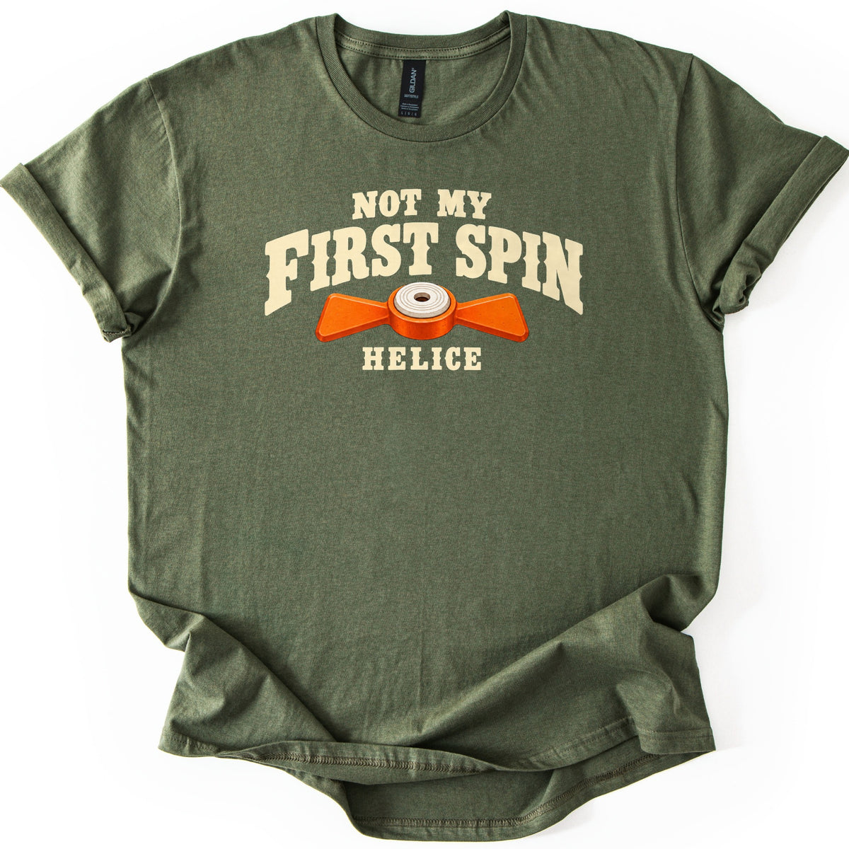 Not My First Spin Comfort Colors T-shirt in Military Green, laid flat. Cream western-style letters read Not My First Spin above an orange two-bladed propeller target with a white center, with the word Helice below. The shirt lies on a white surface with sleeves cuffed.