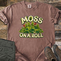Moss on a Roll Comfort Colors T-shirt in Espresso, laid flat. Swirled paper-quill style green moss with two rust-colored sprouts sits between the words Moss On A Roll in cream lettering. Sleeves are cuffed on a wood plank floor beside olive hiking pants and brown hiking boots.