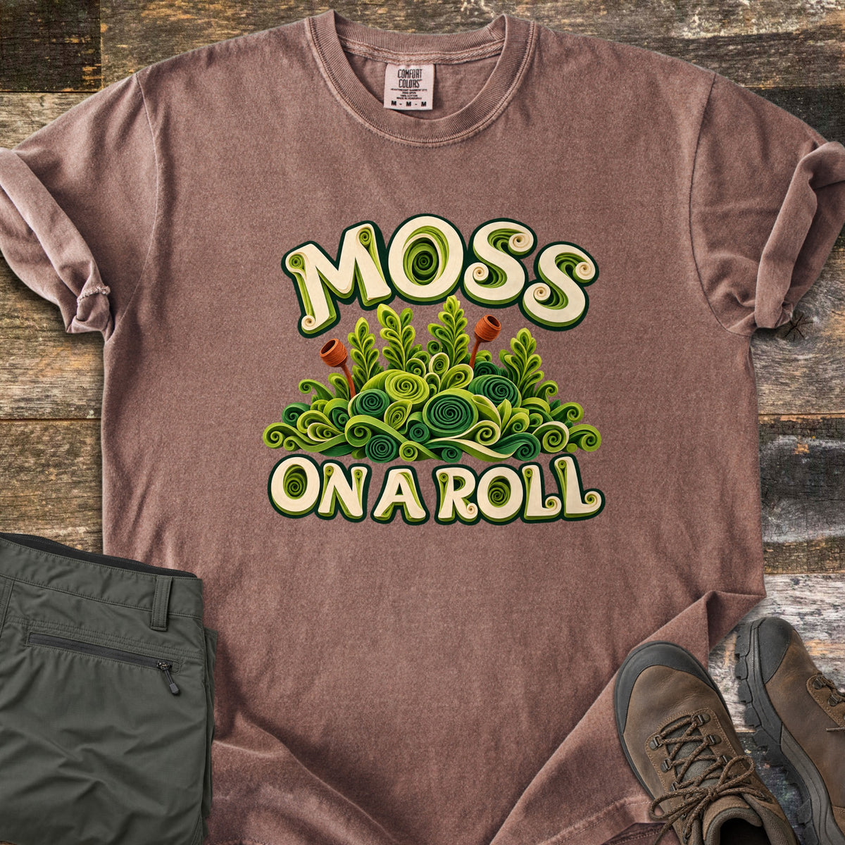 Moss on a Roll Comfort Colors T-shirt in Espresso, laid flat. Swirled paper-quill style green moss with two rust-colored sprouts sits between the words Moss On A Roll in cream lettering. Sleeves are cuffed on a wood plank floor beside olive hiking pants and brown hiking boots.