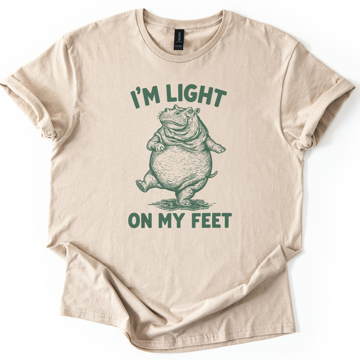 I'm Light On My Feet Comfort Colors T-shirt in Sand, laid flat. A green etched-style drawing of a round hippo standing upright on one foot, mid-step, between the words I'M LIGHT above and ON MY FEET below. The shirt has rolled sleeves on a white background.