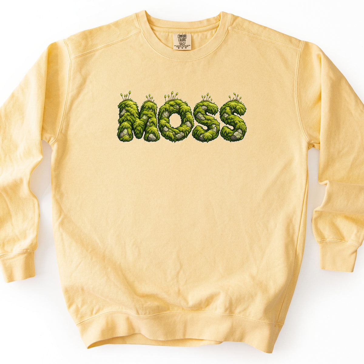 Moss Comfort Colors crewneck sweatshirt in Butter, laid flat. The word MOSS is printed across the chest in large letters made of green moss and gray stones, with small sprouts on top. The sweatshirt lies on a white background.
