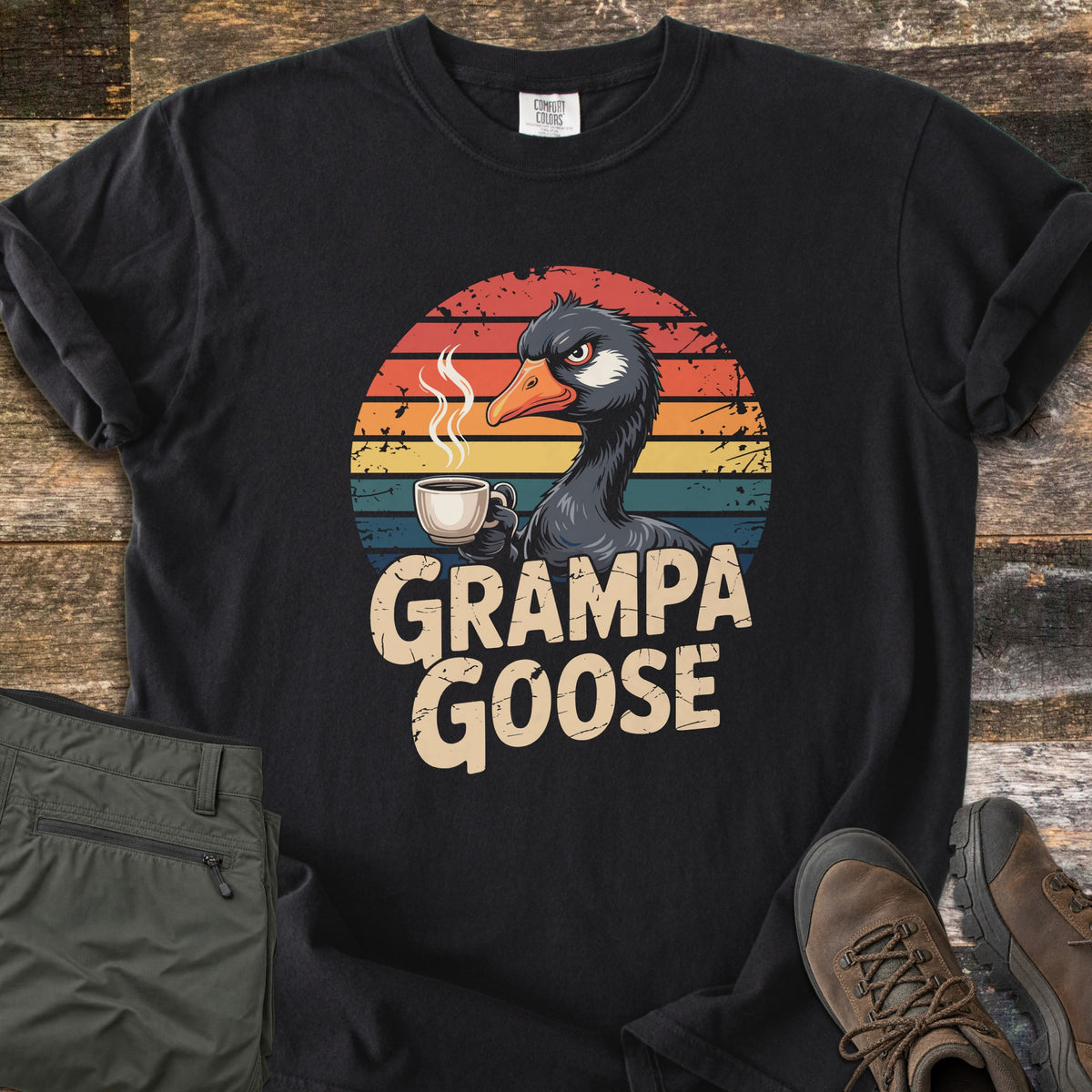 Grampa Goose Comfort Colors T-shirt in Black, laid flat. A grumpy cartoon goose holding a steaming coffee mug in front of a distressed retro sunset of striped bands, above the words Grampa Goose. Olive pants and brown hiking boots sit beside it on weathered wood.